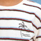 A child wearing the Boys’ Livo T-Shirt by Bonton, a kids’ cotton t-shirt with colored stripes and an embroidered palm tree, sun, and waves design on the chest.