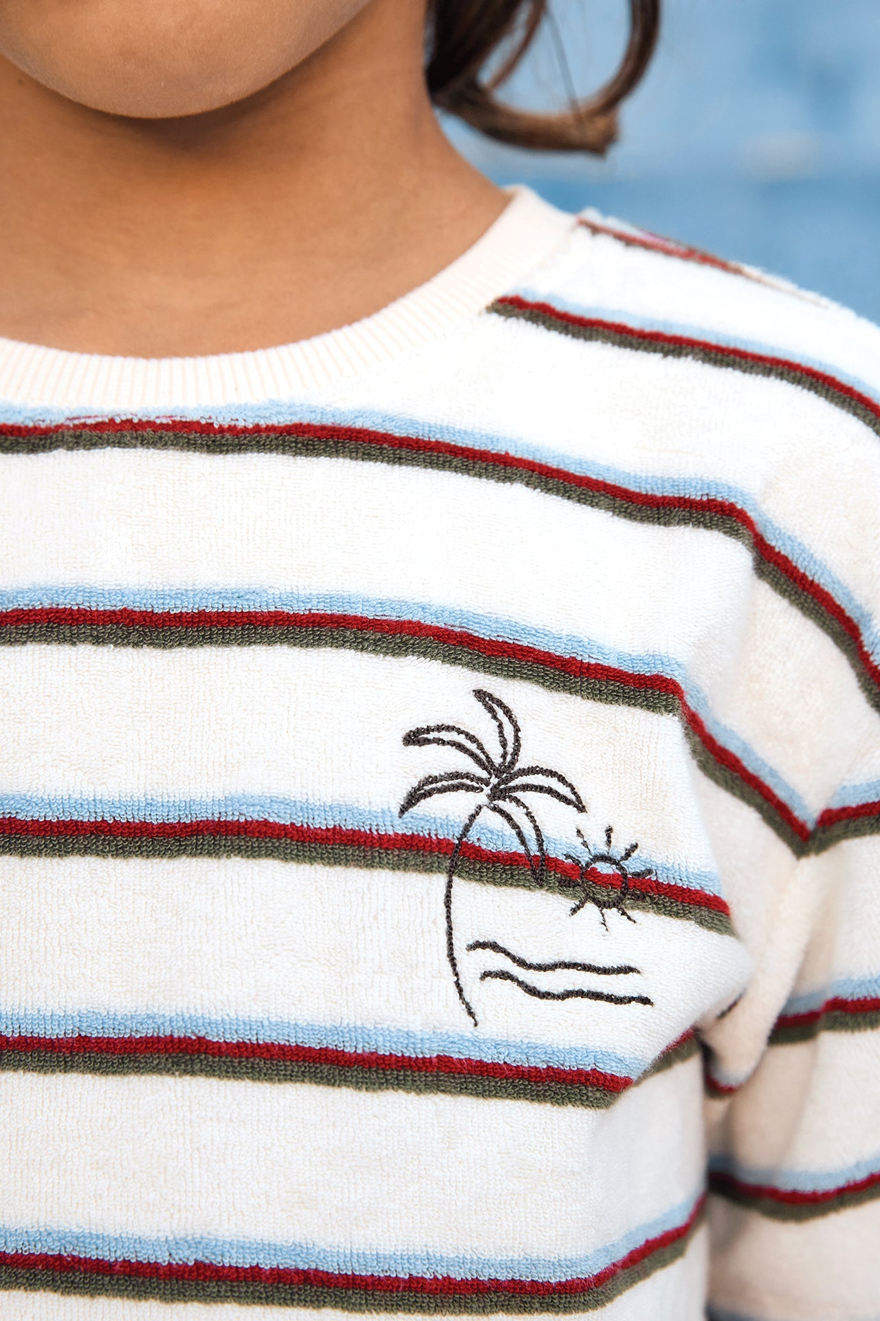A child wearing the Boys’ Livo T-Shirt by Bonton, a kids’ cotton t-shirt with colored stripes and an embroidered palm tree, sun, and waves design on the chest.