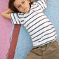 A young child with light brown hair lies on a multicolored cracked surface, wearing the Boys’ Livo T-Shirt by Bonton—an easygoing kids’ cotton tee with subtle embroidered palm tree detailing—paired with khaki pants, arms behind head and a neutral expression.