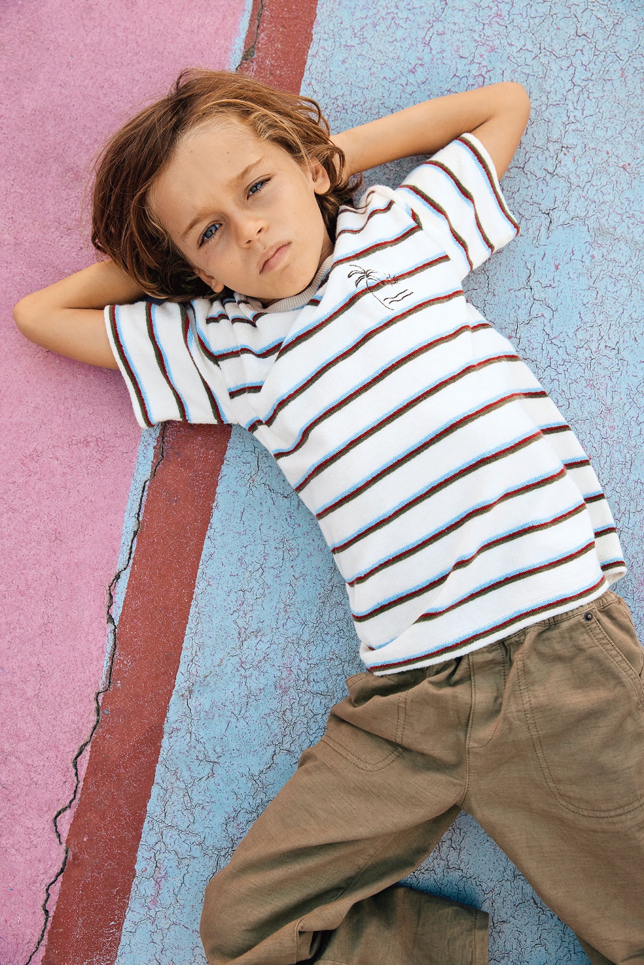A young child with light brown hair lies on a multicolored cracked surface, wearing the Boys’ Livo T-Shirt by Bonton—an easygoing kids’ cotton tee with subtle embroidered palm tree detailing—paired with khaki pants, arms behind head and a neutral expression.