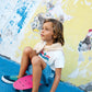 A young child sits on a pink skateboard by a colorful, weathered wall, wearing a white T-shirt, teal sneakers, and Bonton Boys' Naples Shorts.
