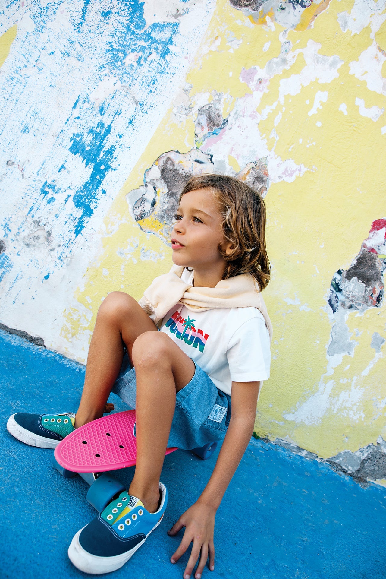 A young child sits on a pink skateboard by a colorful, weathered wall, wearing a white T-shirt, teal sneakers, and Bonton Boys' Naples Shorts.
