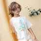 A young child with shoulder-length hair stands against a beige wall, wearing a Bonton Boys' Big Wave Livo T-Shirt with a surfing graphic and matching shorts.