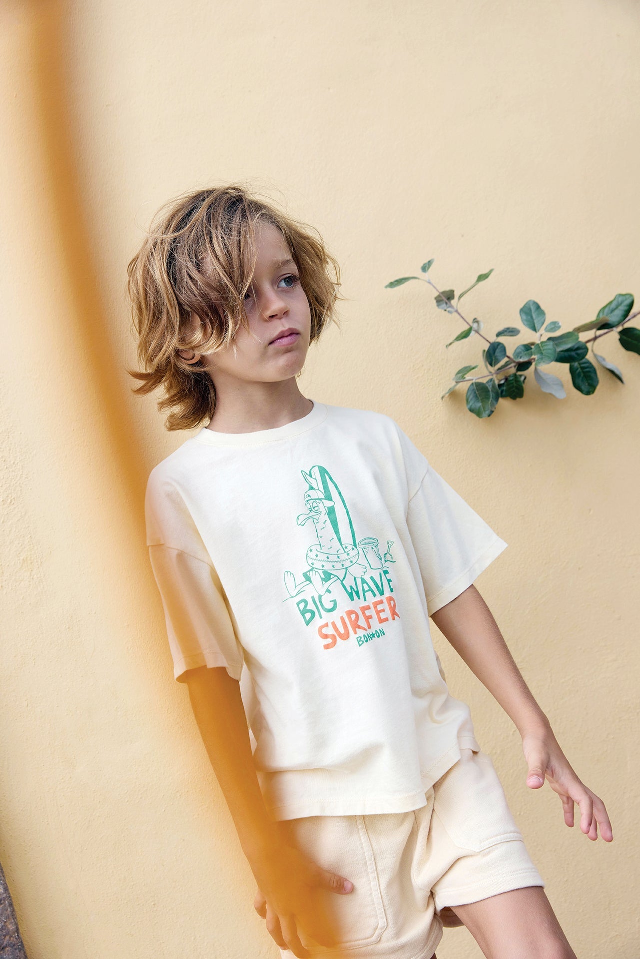 A young child with shoulder-length hair stands against a beige wall, wearing a Bonton Boys' Big Wave Livo T-Shirt with a surfing graphic and matching shorts.