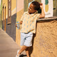 A young boy in a Bonton Boys' Livio Polo, Palmiers, leans against a wall outside a building, holding a drink near a “Frutería” sign with a pineapple illustration.