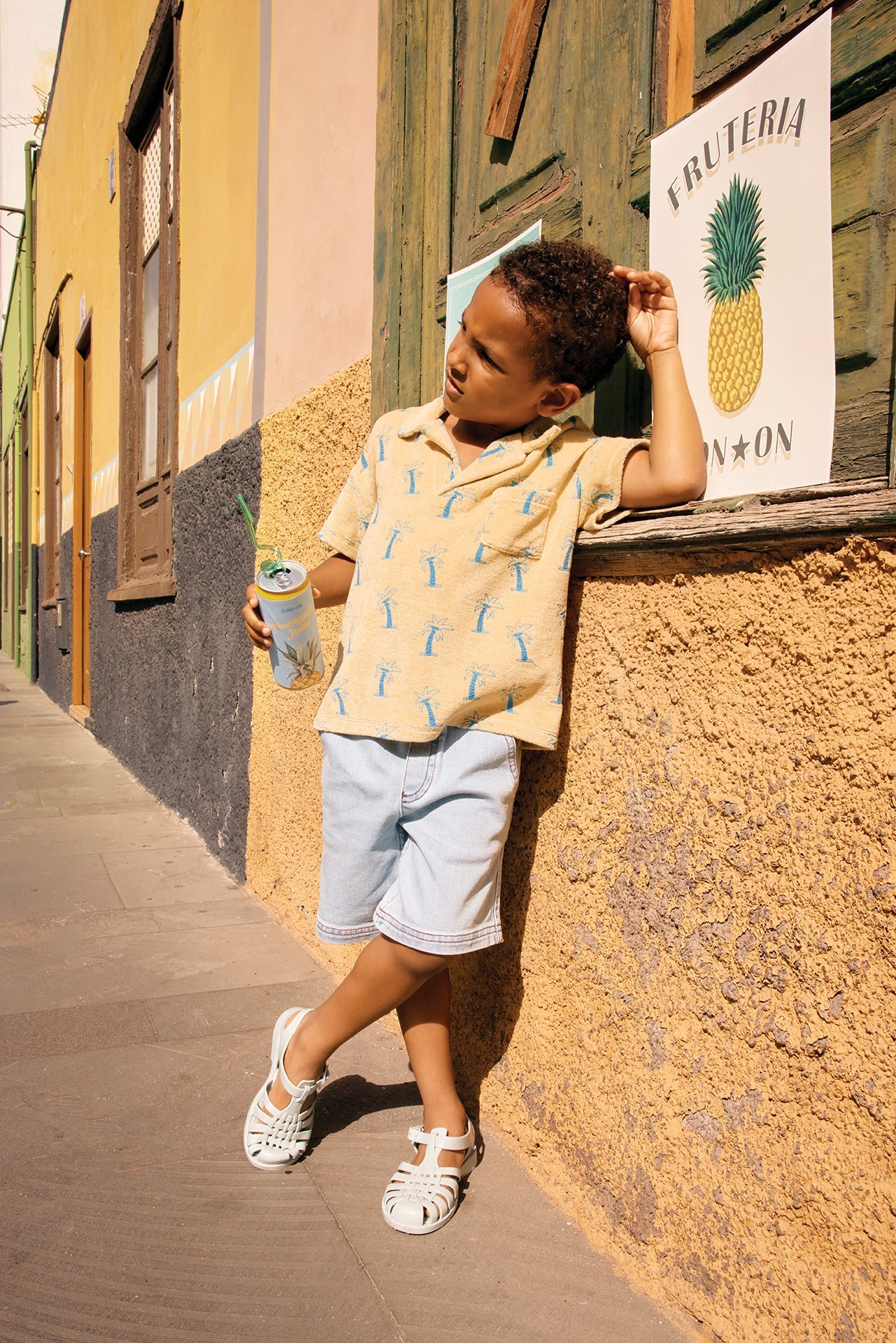 A young boy in a Bonton Boys' Livio Polo, Palmiers, leans against a wall outside a building, holding a drink near a “Frutería” sign with a pineapple illustration.