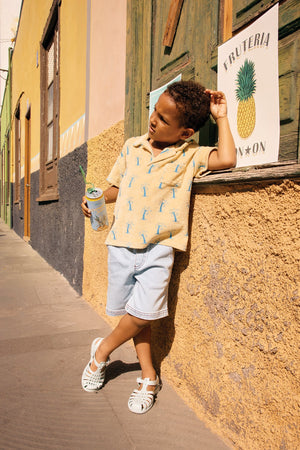 A young boy in a Bonton Boys' Livio Polo, Palmiers, leans against a wall outside a building, holding a drink near a “Frutería” sign with a pineapple illustration.