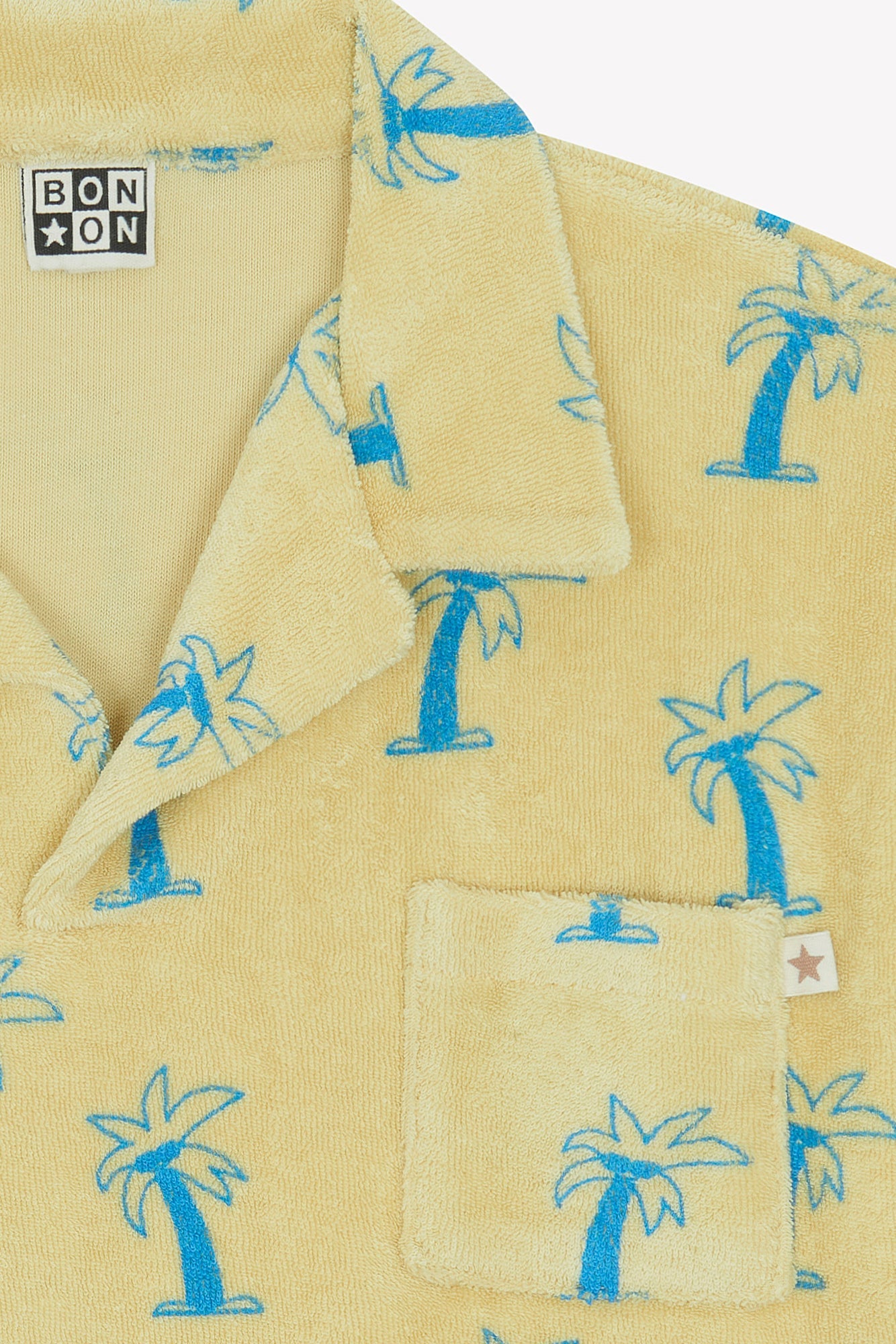 Close-up of the Boys' Livio Polo, Palmiers by Bonton—a yellow 100% cotton terry polo with blue palm tree prints, a patch pocket with star tag, and a Bonton label inside the collar.