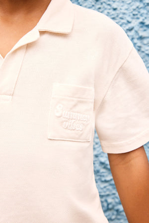 A boy wearing Bonton’s Boys' Livio Polo—a cream-colored cotton shirt with a "Summer Vibes" pocket embroidery—stands against a blue textured wall, perfectly capturing an effortless summer style.