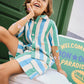 A smiling child wearing the Bonton Boys’ Lois Short—blue, green, and white striped cotton shorts with a matching top—sits on a cooler, colorful stickers on their arms, in front of a green door and a bright welcome sign.