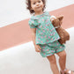 A young child stands outdoors with a stuffed animal, wearing the Bonton Baby Girls' Aria Shirt featuring a green floral pattern and Peter Pan collar, paired with pink sandals and a neutral expression.