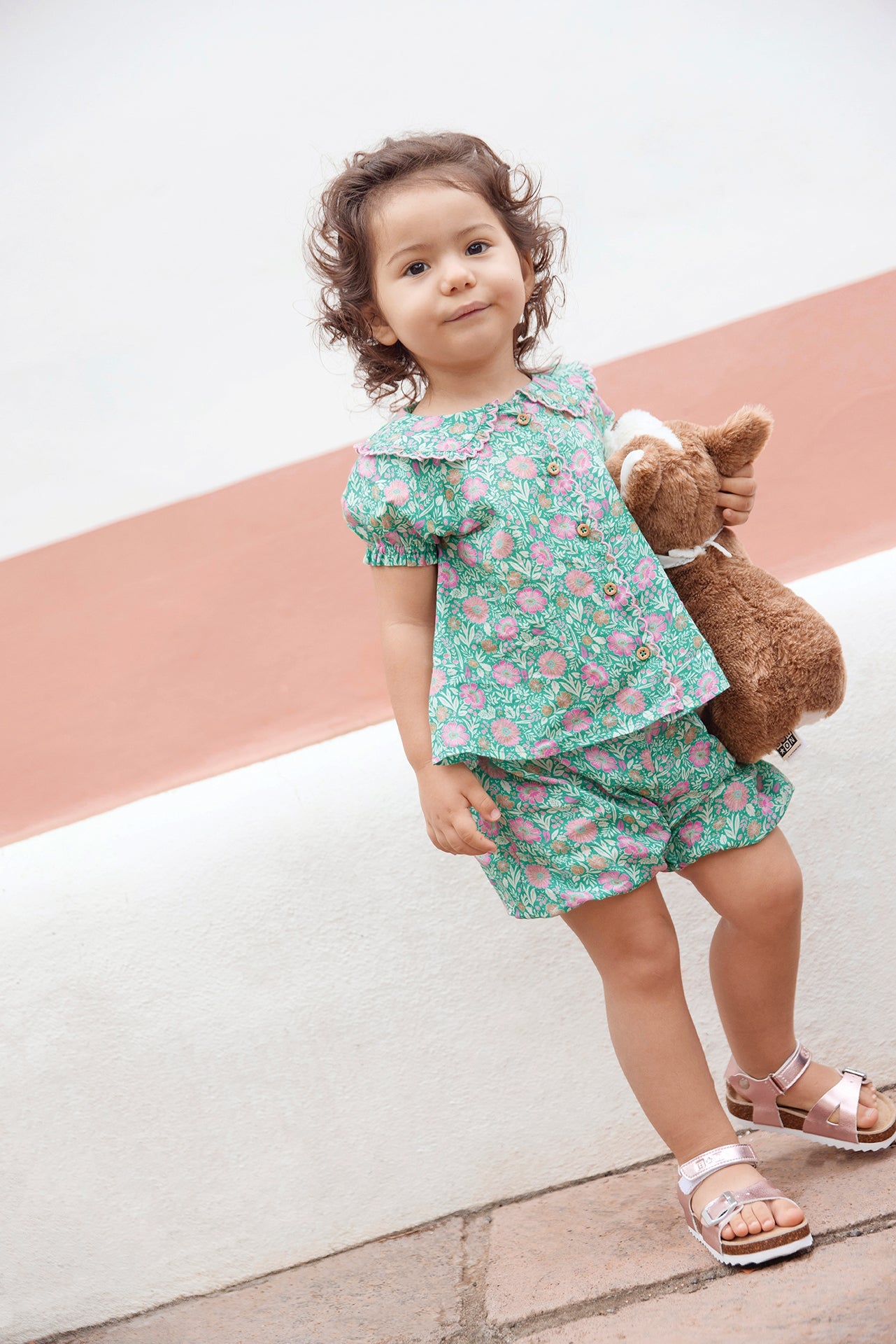 A young child stands outdoors with a stuffed animal, wearing the Bonton Baby Girls' Aria Shirt featuring a green floral pattern and Peter Pan collar, paired with pink sandals and a neutral expression.