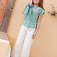 A young girl stands by a peach wall in a green floral top, pink shoes, and Bonton’s Girls' Lakiko Pant—white wide-cut with an embroidered pocket—gently touching her face and smiling softly.