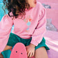 A young child with curly hair, dressed in a Bonton Girls’ Nolwen Sweatshirt with puffy sleeves and green shorts, sits on pastel pink steps holding a pink skateboard and a frozen drink, looking to the side with mouth open.