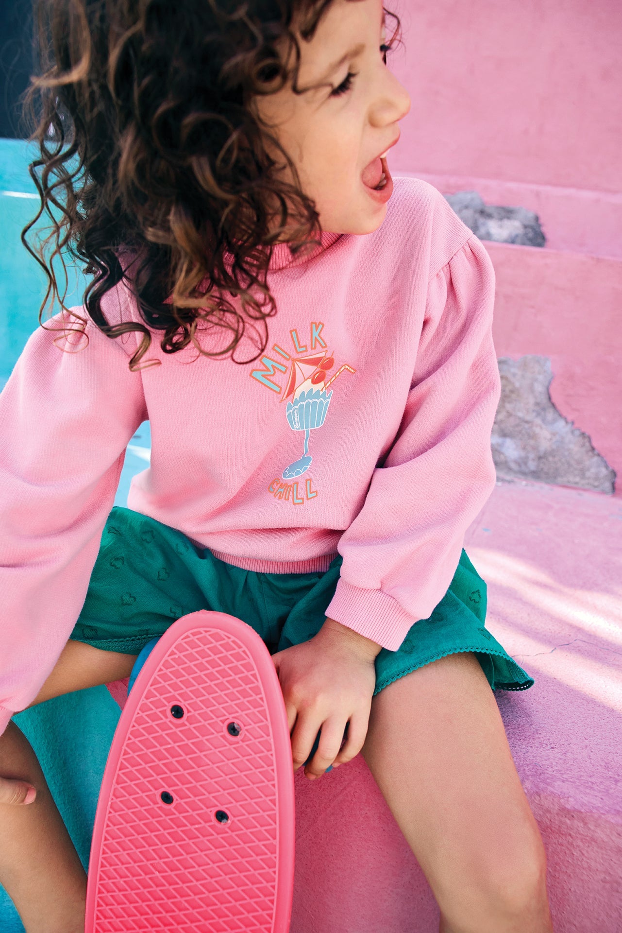 A young child with curly hair, dressed in a Bonton Girls’ Nolwen Sweatshirt with puffy sleeves and green shorts, sits on pastel pink steps holding a pink skateboard and a frozen drink, looking to the side with mouth open.