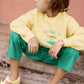 A child sits on a stone step, wearing Bonton's Boys' Paradise Sweatshirt—ultra-soft cotton—paired with green pants and yellow sandals, looking to the side with a neutral expression against a pink wall.