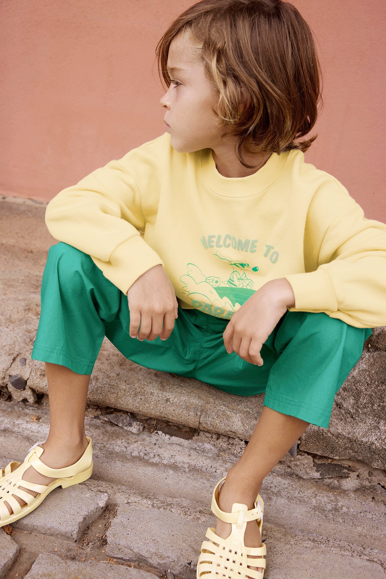A child sits on a stone step, wearing Bonton's Boys' Paradise Sweatshirt—ultra-soft cotton—paired with green pants and yellow sandals, looking to the side with a neutral expression against a pink wall.