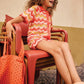 A young girl with curly hair sits on red chairs, wearing a Bonton Girls' Ninon Knit Top made from 100% cotton in a red and white pattern, with an orange bag by her side.