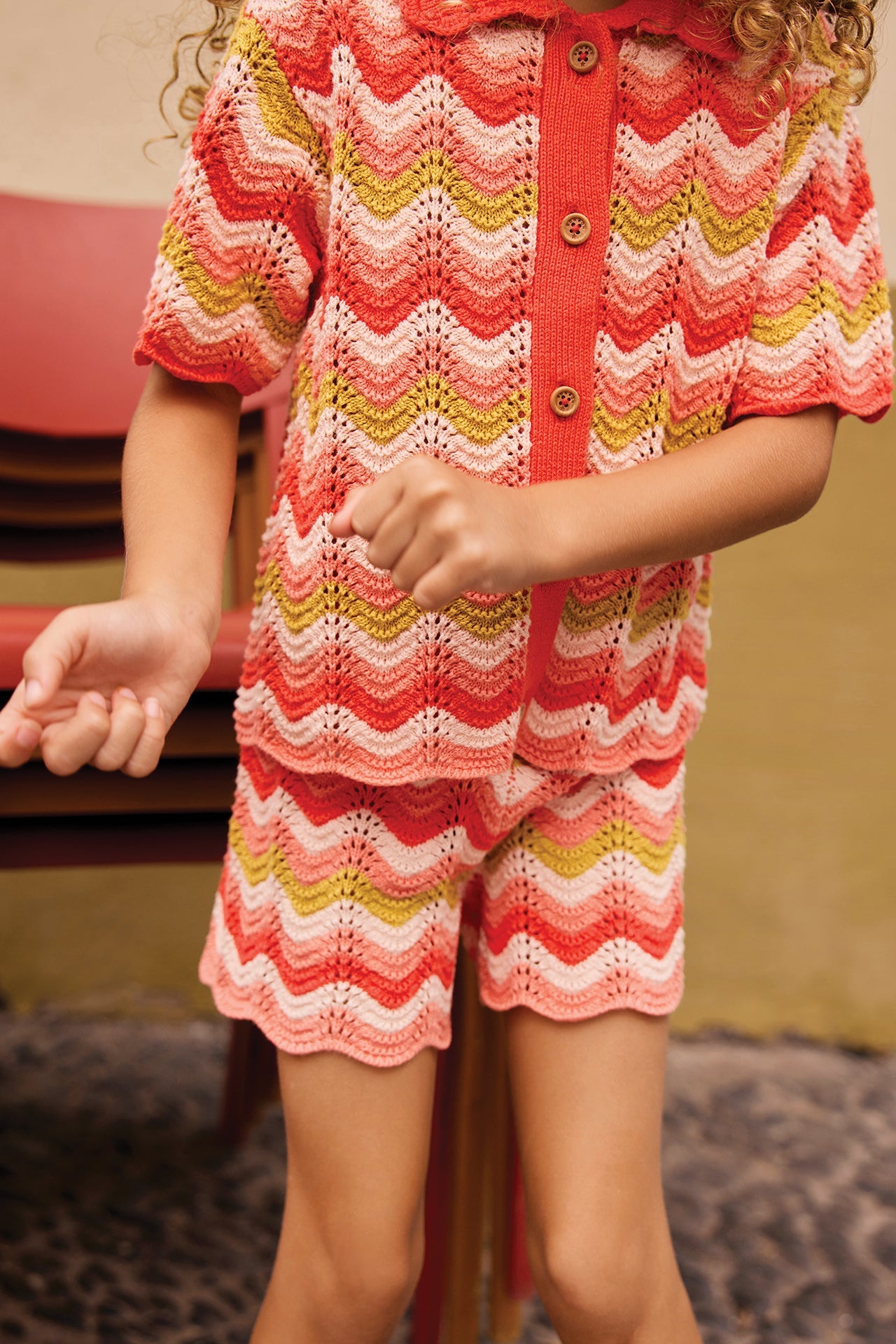 A child in Bonton Girls' Nine Shorts and a colorful, herringbone-patterned knit shirt stands beside a stack of red chairs on cobblestones.