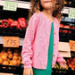 A young girl with curly hair wears the Bonton Girls' Lilou Cardigan with openwork hearts over a green dress, standing in front of a market stall filled with fruits, vegetables, and handwritten price signs.