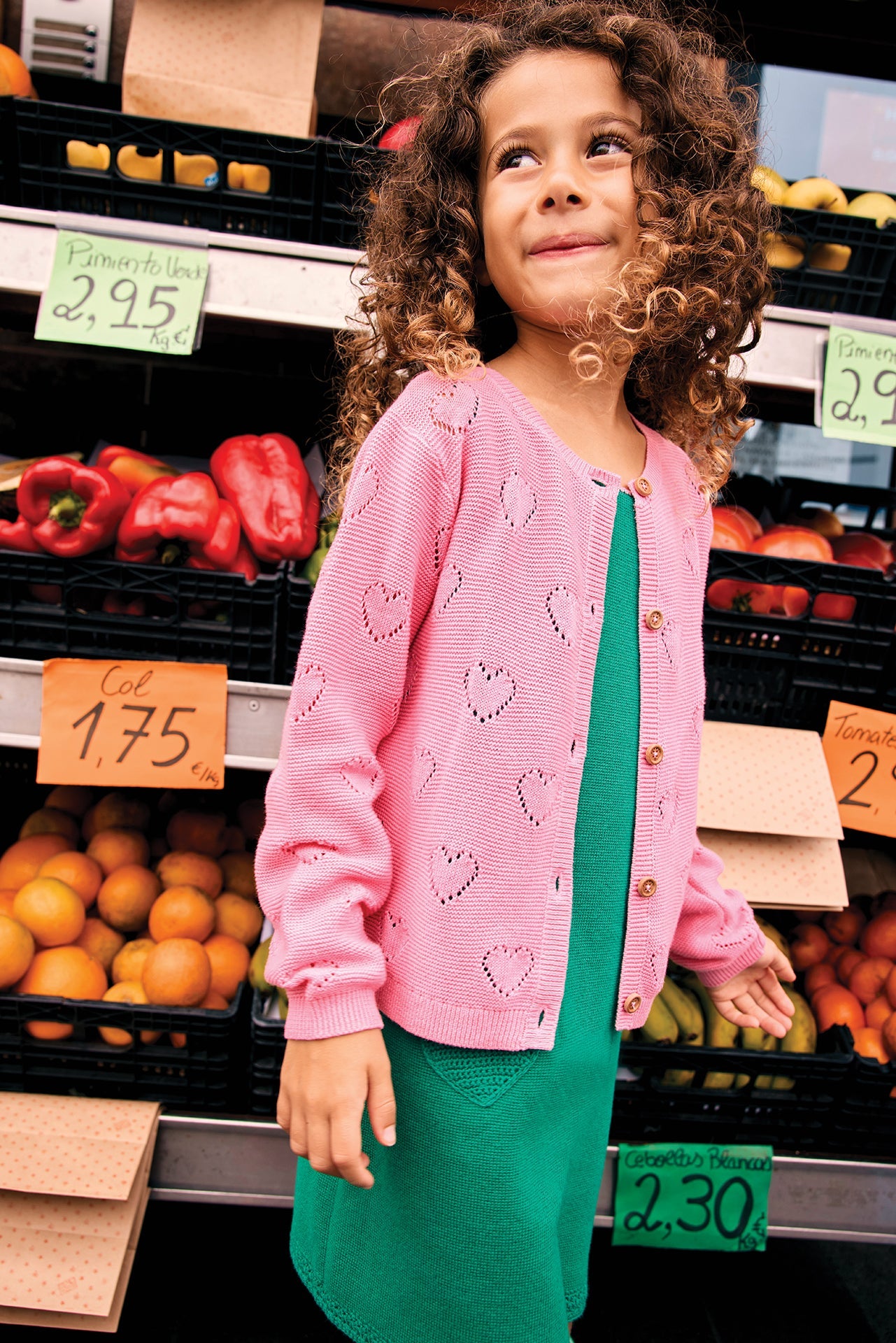 A young girl with curly hair wears the Bonton Girls' Lilou Cardigan with openwork hearts over a green dress, standing in front of a market stall filled with fruits, vegetables, and handwritten price signs.