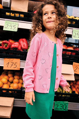 A young girl with curly hair wears the Bonton Girls' Lilou Cardigan with openwork hearts over a green dress, standing in front of a market stall filled with fruits, vegetables, and handwritten price signs.