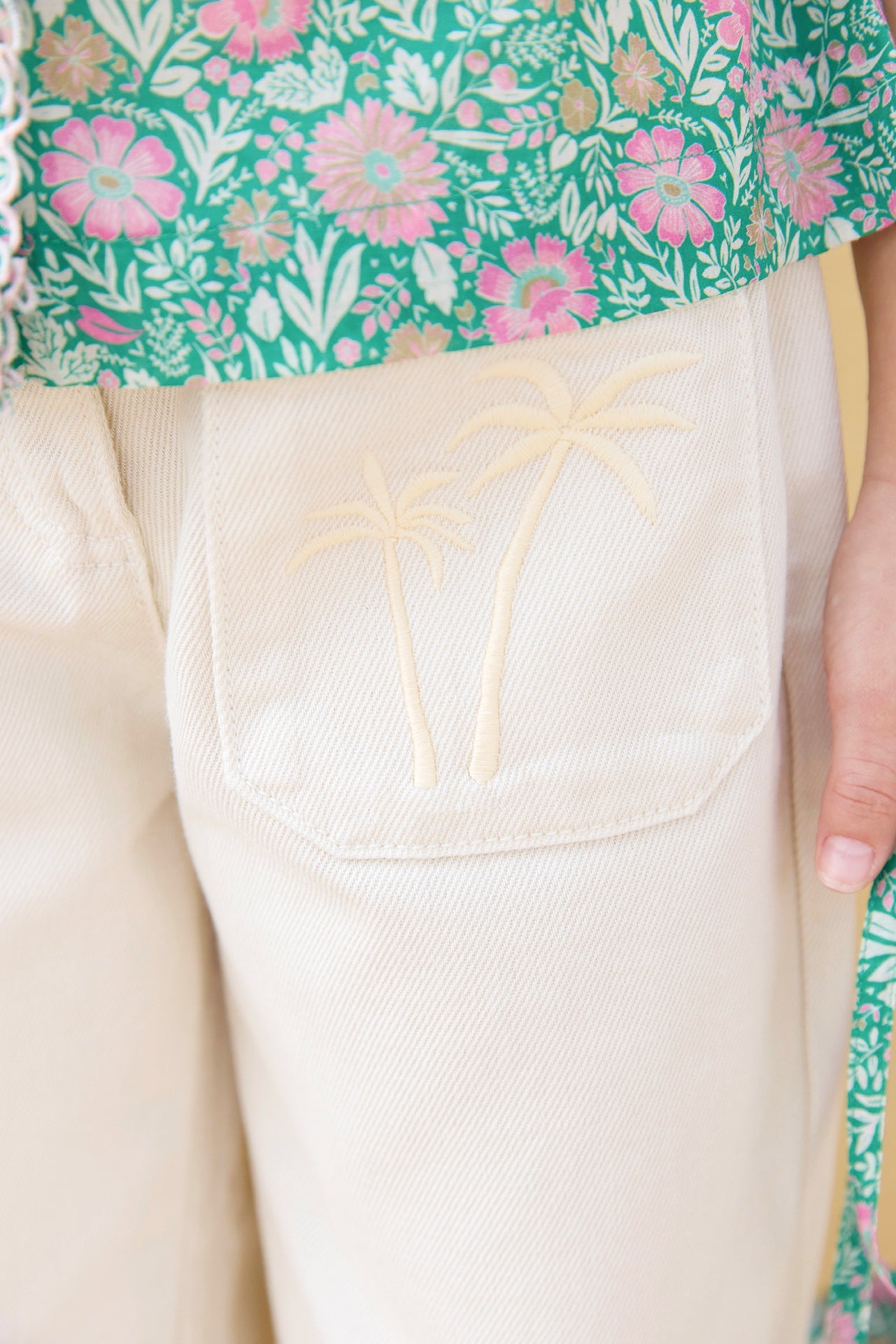 Close-up of Bonton Girls' Lakiko Pant in cream, featuring a wide cut and an embroidered palm tree pocket, styled with a green floral-patterned top for a playful look.