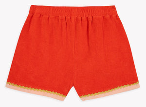 The Girls’ Livi Short by Bonton showcases bright red soft cotton terry cloth, viewed from the back, with an elastic waistband and pink trim accented by green wavy crochet detailing along the bottom hems.