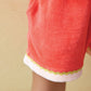 A close-up of a child wearing the Bonton Girls’ Livi Short—bright coral with white and yellow trim, crafted from soft cotton and featuring an elastic waist—standing against a beige background, perfect for warm weather play.