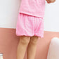 A young child stands against a pink and white wall, wearing a pink t-shirt, Bonton Baby Girls' Bombe Bloomer, and metallic sandals. A white bottle with colored dots lies on the ground nearby.