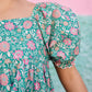 A child models the Bonton Girls' Georgette Dress in green with pink floral patterns, set against a pink and green background.