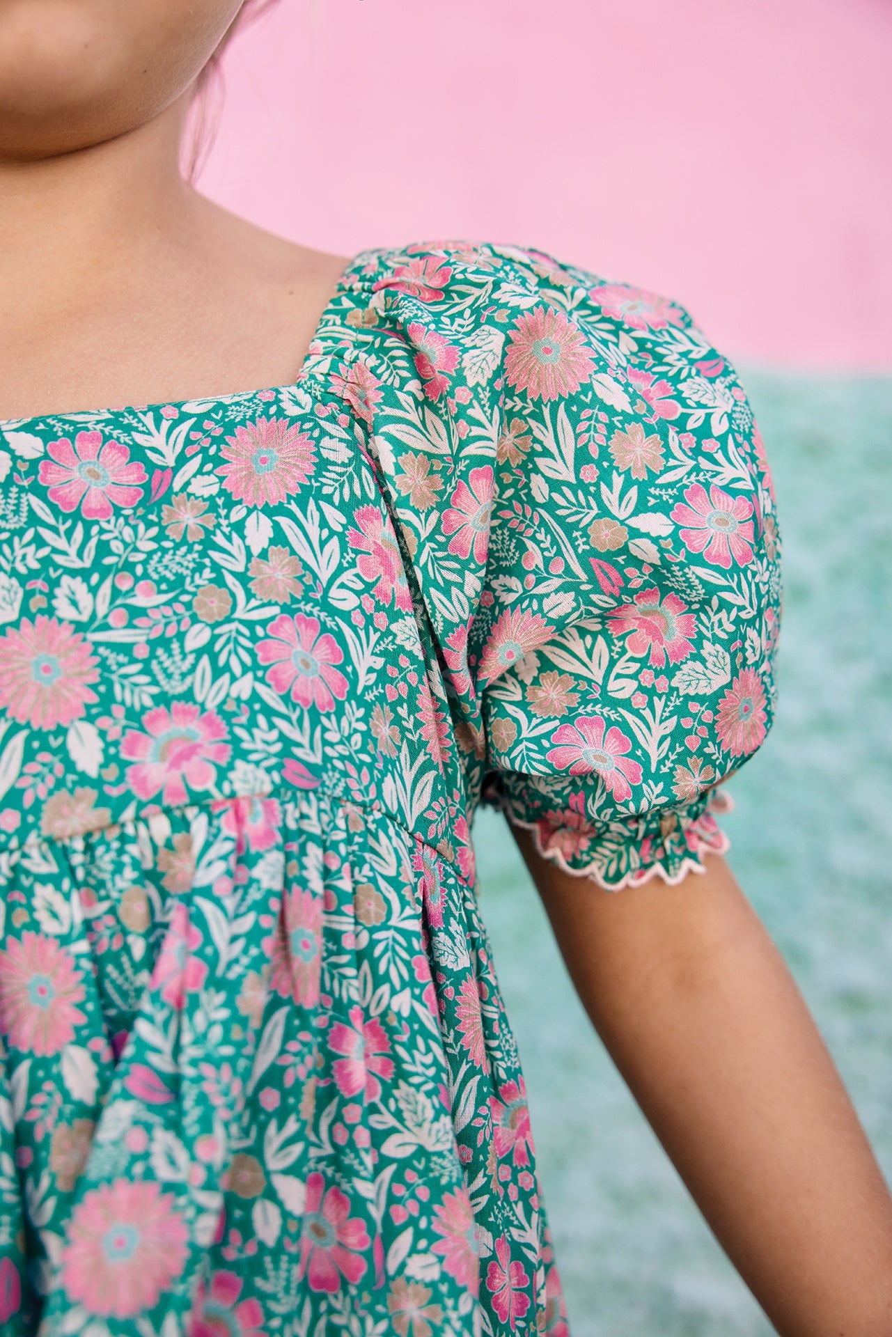 A child models the Bonton Girls' Georgette Dress in green with pink floral patterns, set against a pink and green background.