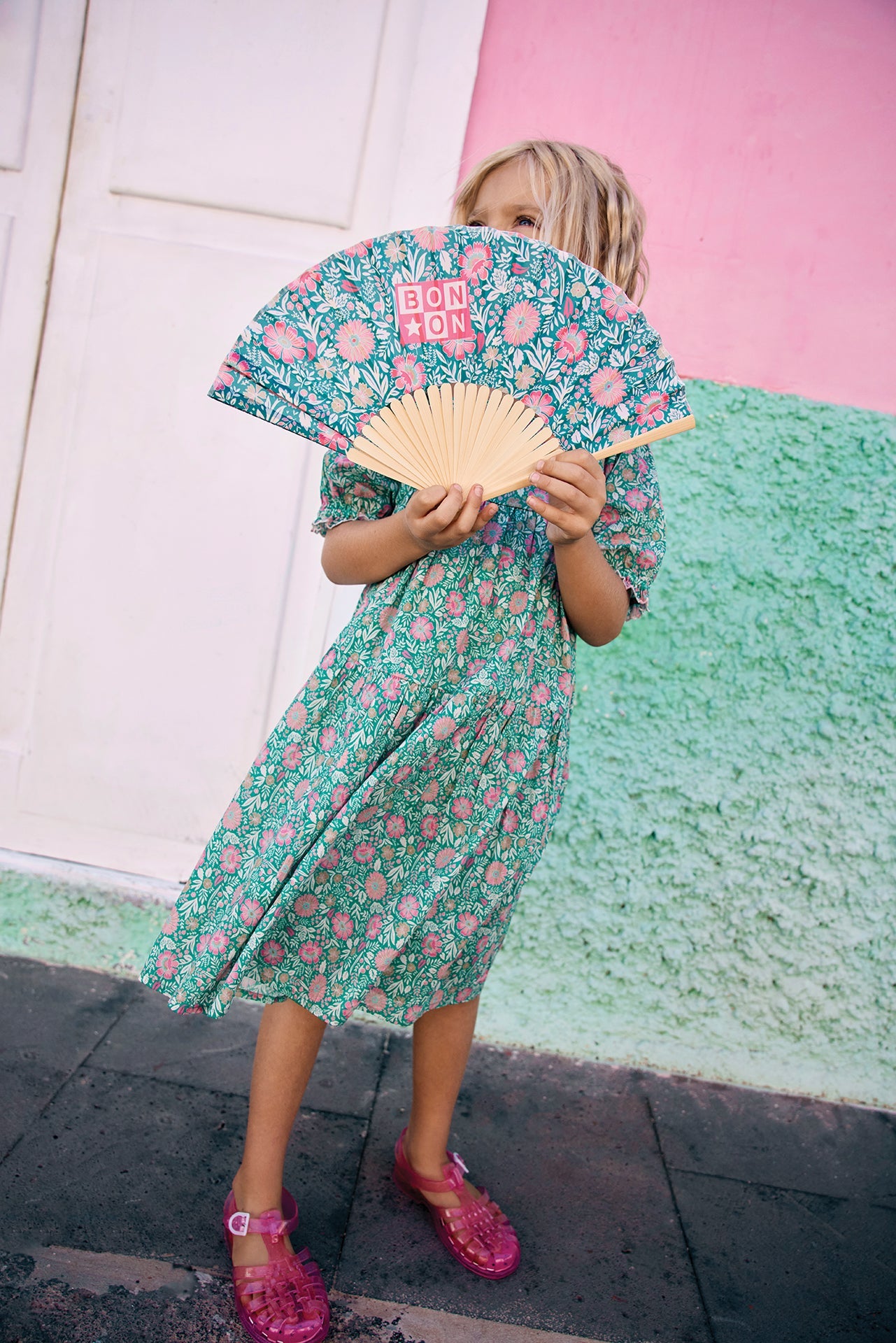 A child in a green floral Bonton Girls' Georgette Dress holds a matching decorative fan, standing before a pink and green textured wall.
