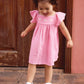 A young child wears the Bonton Baby Girls' Revebb Dress and silver sandals, standing on a stone step by a wooden door and looking down with a slight smile.