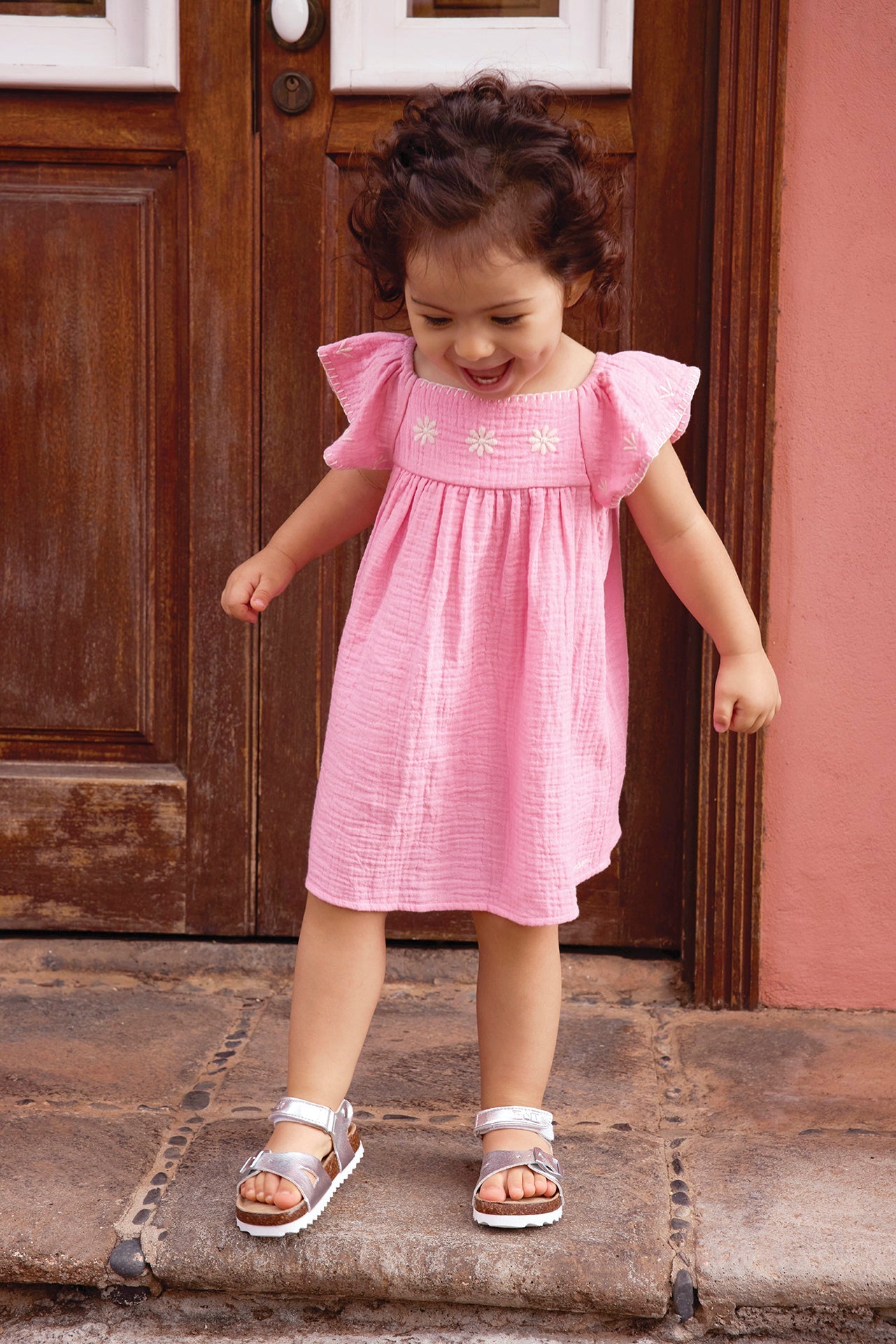 A young child wears the Bonton Baby Girls' Revebb Dress and silver sandals, standing on a stone step by a wooden door and looking down with a slight smile.