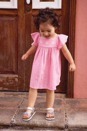 A young child wears the Bonton Baby Girls' Revebb Dress and silver sandals, standing on a stone step by a wooden door and looking down with a slight smile.