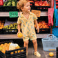 A young child wearing the Bonton Baby Boys’ Lilo Onesie stands among crates of produce at an outdoor market, holding a yellow fruit, with tomatoes, peppers, and bananas in the background for a breezy tropical look.