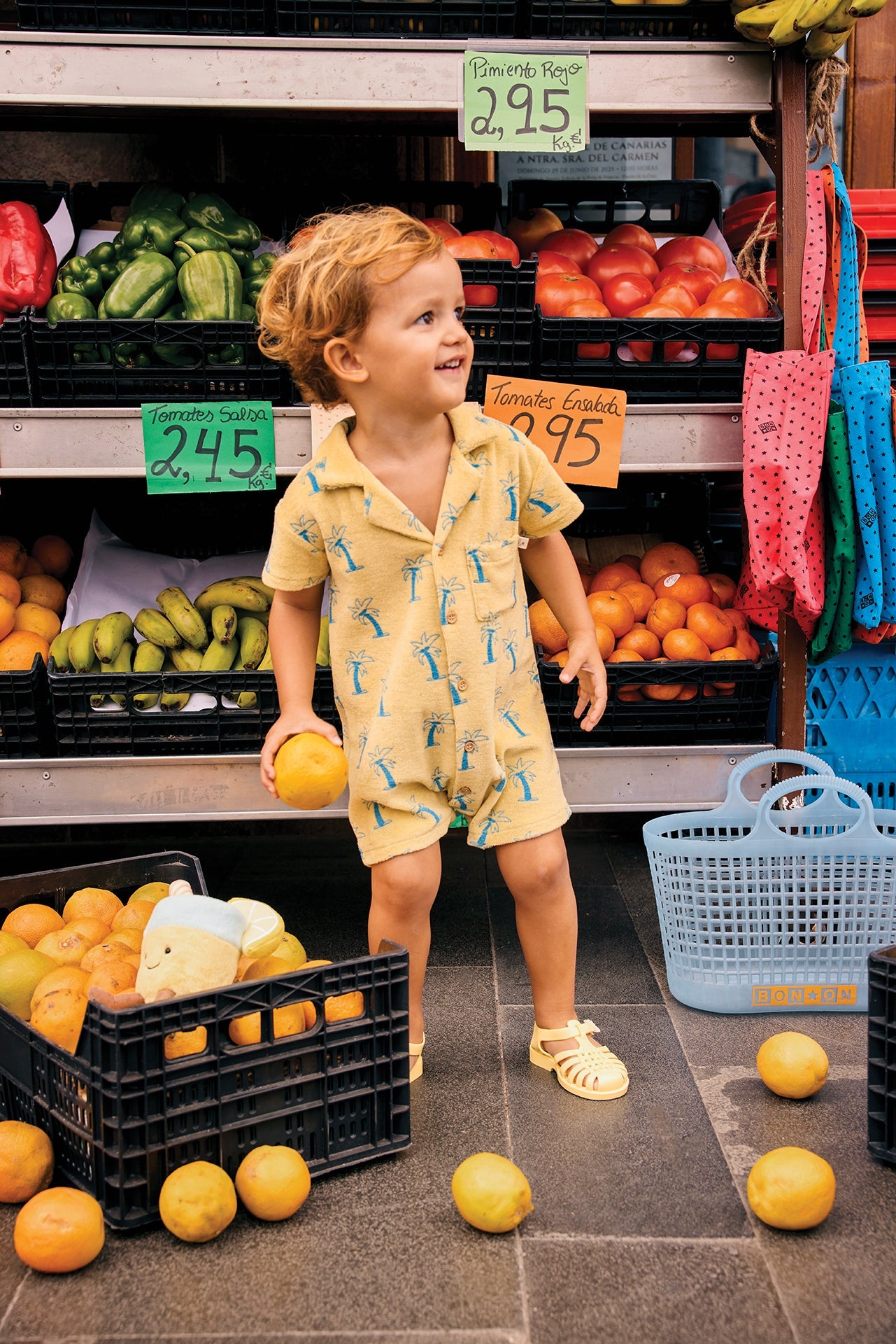 A young child wearing the Bonton Baby Boys’ Lilo Onesie stands among crates of produce at an outdoor market, holding a yellow fruit, with tomatoes, peppers, and bananas in the background for a breezy tropical look.
