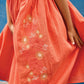 A girl wears the coral-orange Girls' Nicky Dress by Bonton, embroidered with flowers, standing against a blue wall while holding and slightly lifting the sides of her dress.