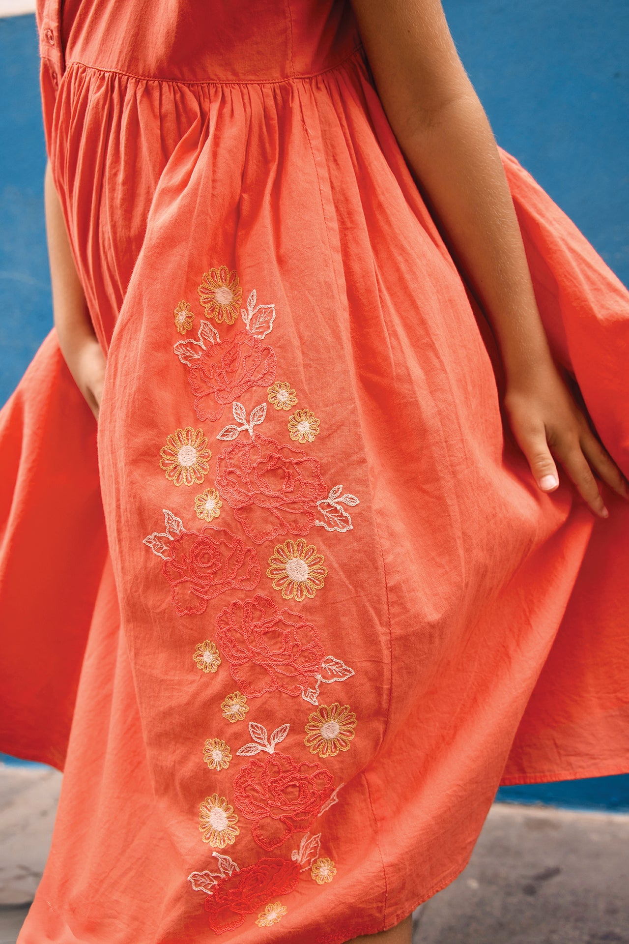 A girl wears the coral-orange Girls' Nicky Dress by Bonton, embroidered with flowers, standing against a blue wall while holding and slightly lifting the sides of her dress.