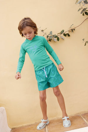 A young child wears the Bonton Kids' Rashguard—a green UV protection swim t-shirt with matching shorts—and white sandals while standing on a tiled floor against a yellow wall decorated with leaves.
