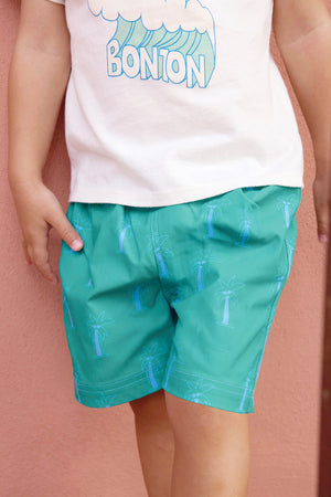 A child in a white Bonton t-shirt and Baby Boys' Ocean Palm Swim Trunk by Bonton, featuring blue palm tree prints, stands against a pink wall, ready for kids beachwear adventures.