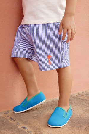 A young child stands against a peach-colored wall in a white shirt, blue striped shorts with an orange logo, and soft cotton Bonton Boys' and Girls' John Slippers for extra comfort.
