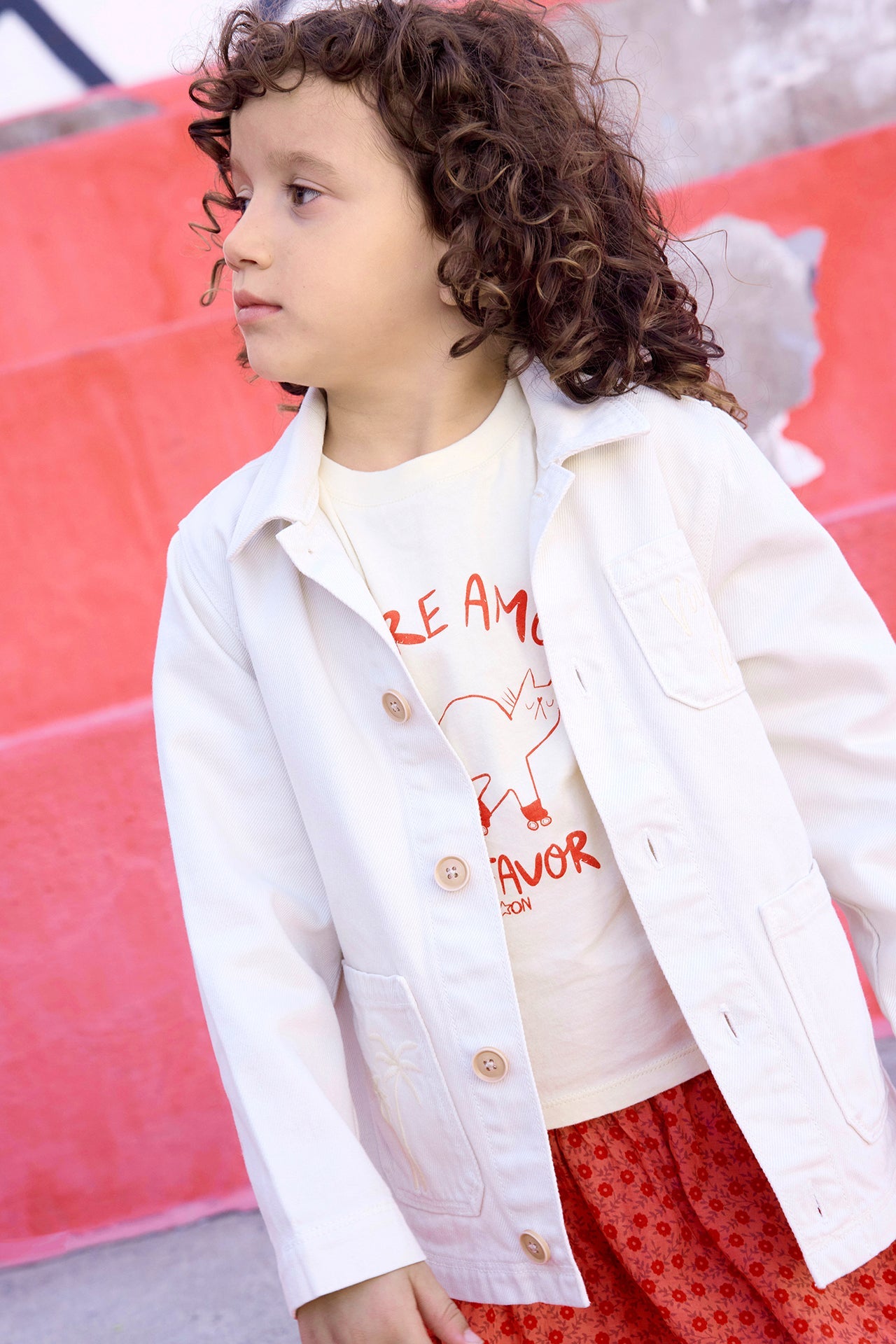 A young child with curly hair stands outdoors, wearing the Bonton Girls' Ermana Jacket over a printed T-shirt and red patterned pants, with red steps in the background.