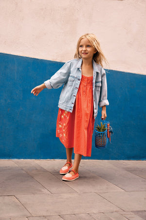 A young girl in the Bonton Girls' Nicky Dress and a denim jacket walks on a paved path, carrying a small basket, in front of a blue and white wall.