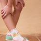 A child stands on a tiled floor wearing pastel green Mary Jane shoes with pink trim and Bonton Girls' Cocofruit Socks featuring playful pineapple designs.