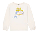 The Bonton Girls’ Mermaid Patch T-Shirt is a white long-sleeve top adorned with “I MISS MERMAID SEASON” text above charming patches of a mermaid tail, shells, and a net bag hanging on a clothesline.