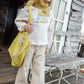 A young child with straight blonde hair stands on a wooden porch, wearing beige pants, a cream long-sleeve shirt layered under the whimsical Bonton Girls’ Mermaid Patch T-Shirt, accessorized with a yellow scarf and holding a yellow bag, next to a rustic wooden bench.