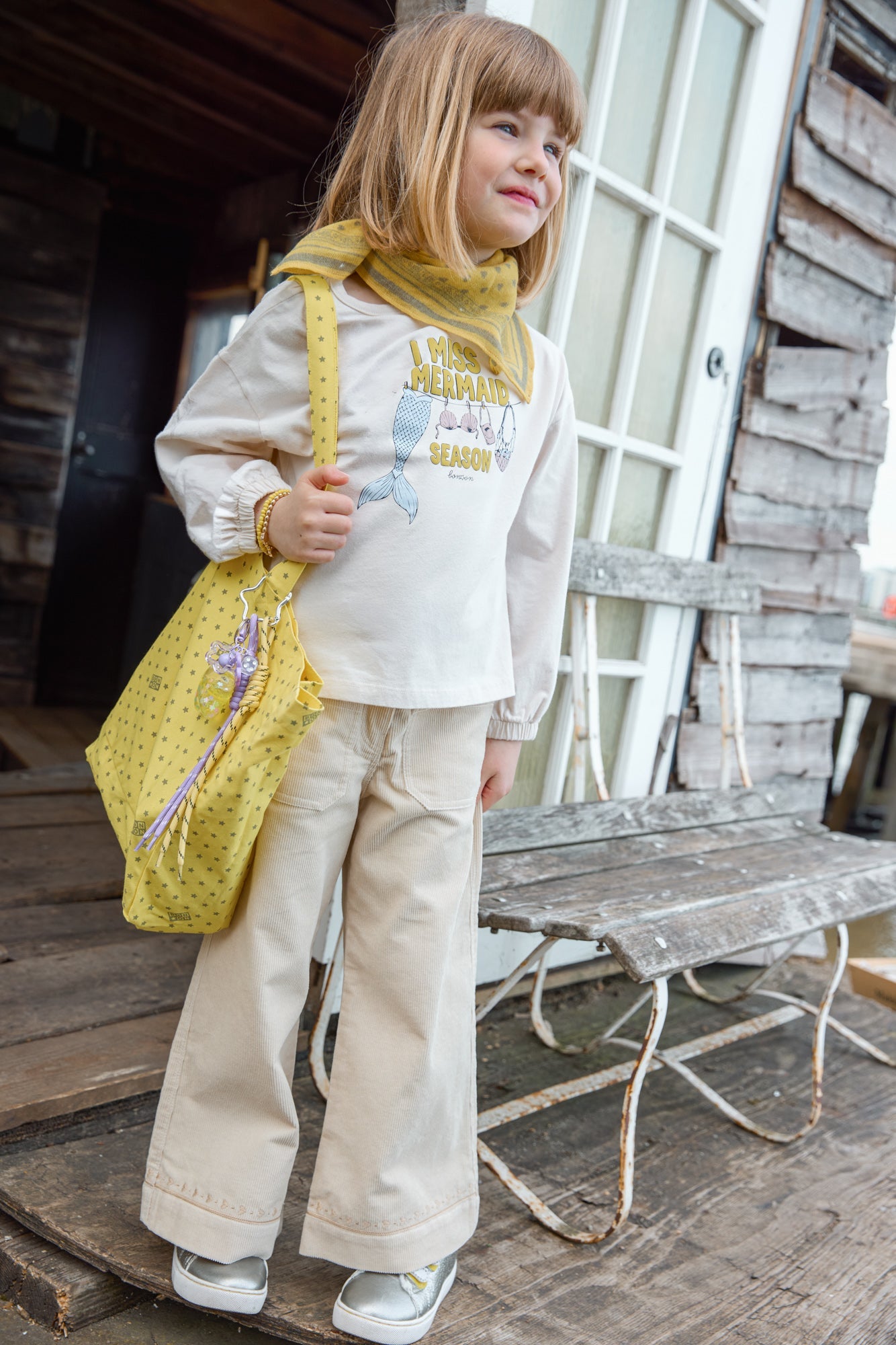 A young child with straight blonde hair stands on a wooden porch, wearing beige pants, a cream long-sleeve shirt layered under the whimsical Bonton Girls’ Mermaid Patch T-Shirt, accessorized with a yellow scarf and holding a yellow bag, next to a rustic wooden bench.