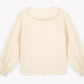 The Girls’ Figue Shirt by Bonton is a long-sleeve cream blouse featuring a delicate Peter Pan collar and ruffled cuffs, crafted from 100% cotton, and displayed flat on a plain white background.